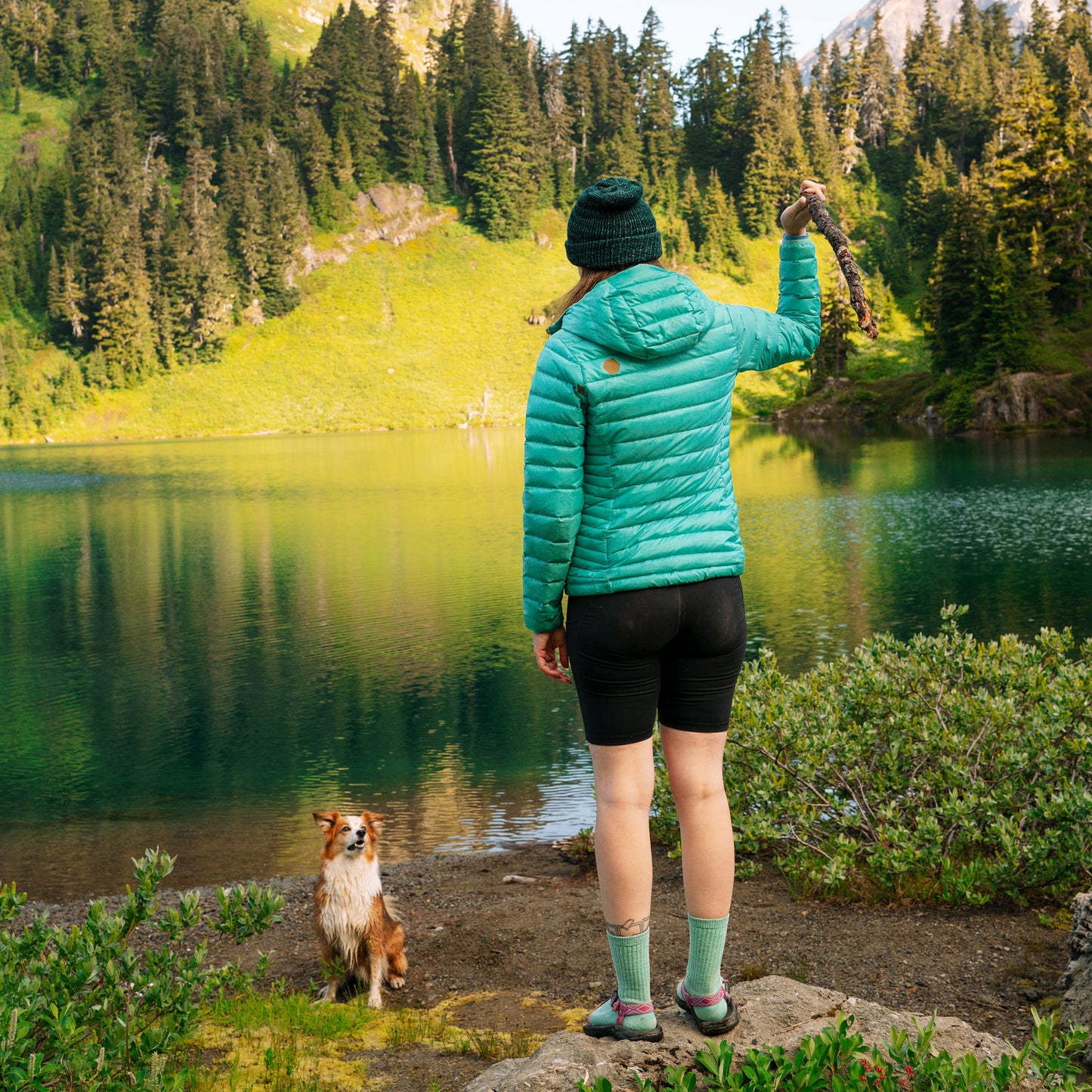 Person in a teal jacket standing by a lake with a dog, surrounded by trees