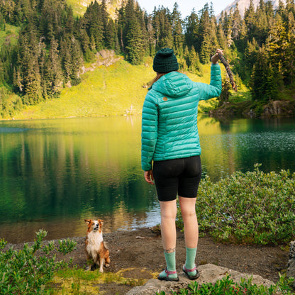 Person in a teal jacket standing by a lake with a dog, surrounded by trees