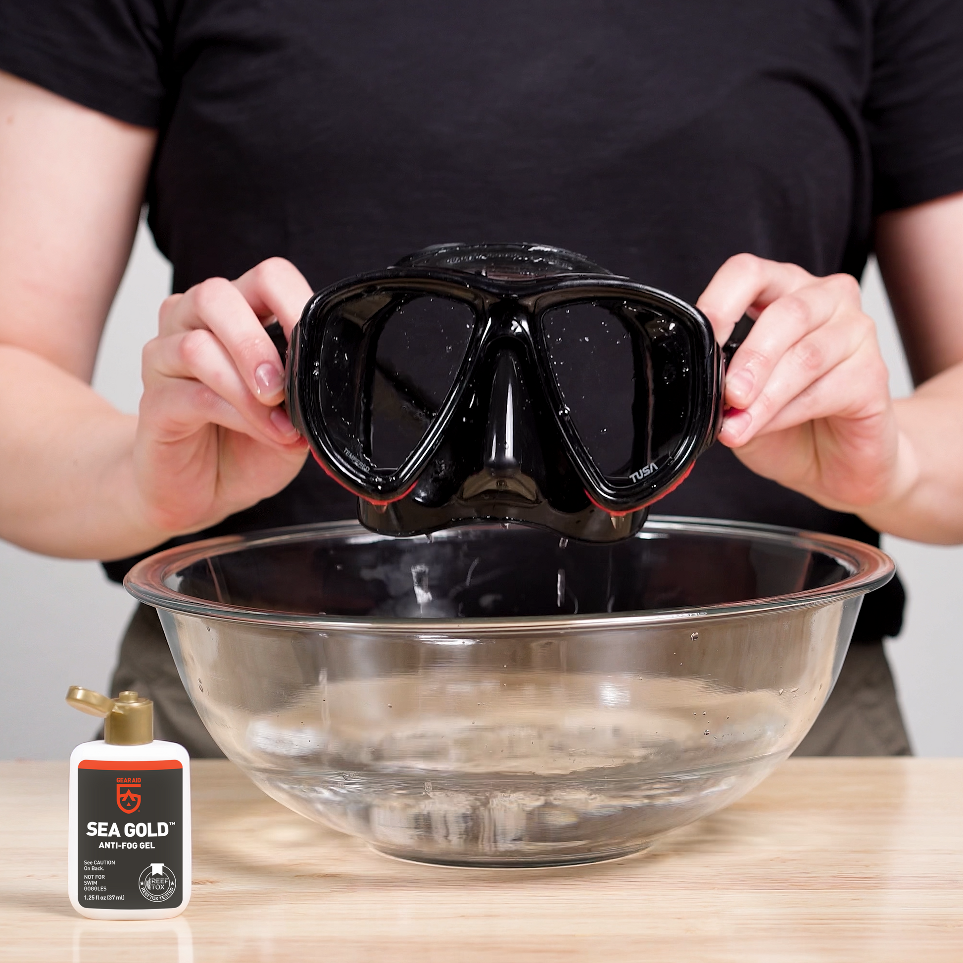 Woman holding a dive mask above a bowl of water next to a bottle of Sea Gold Anti-Fog Gel