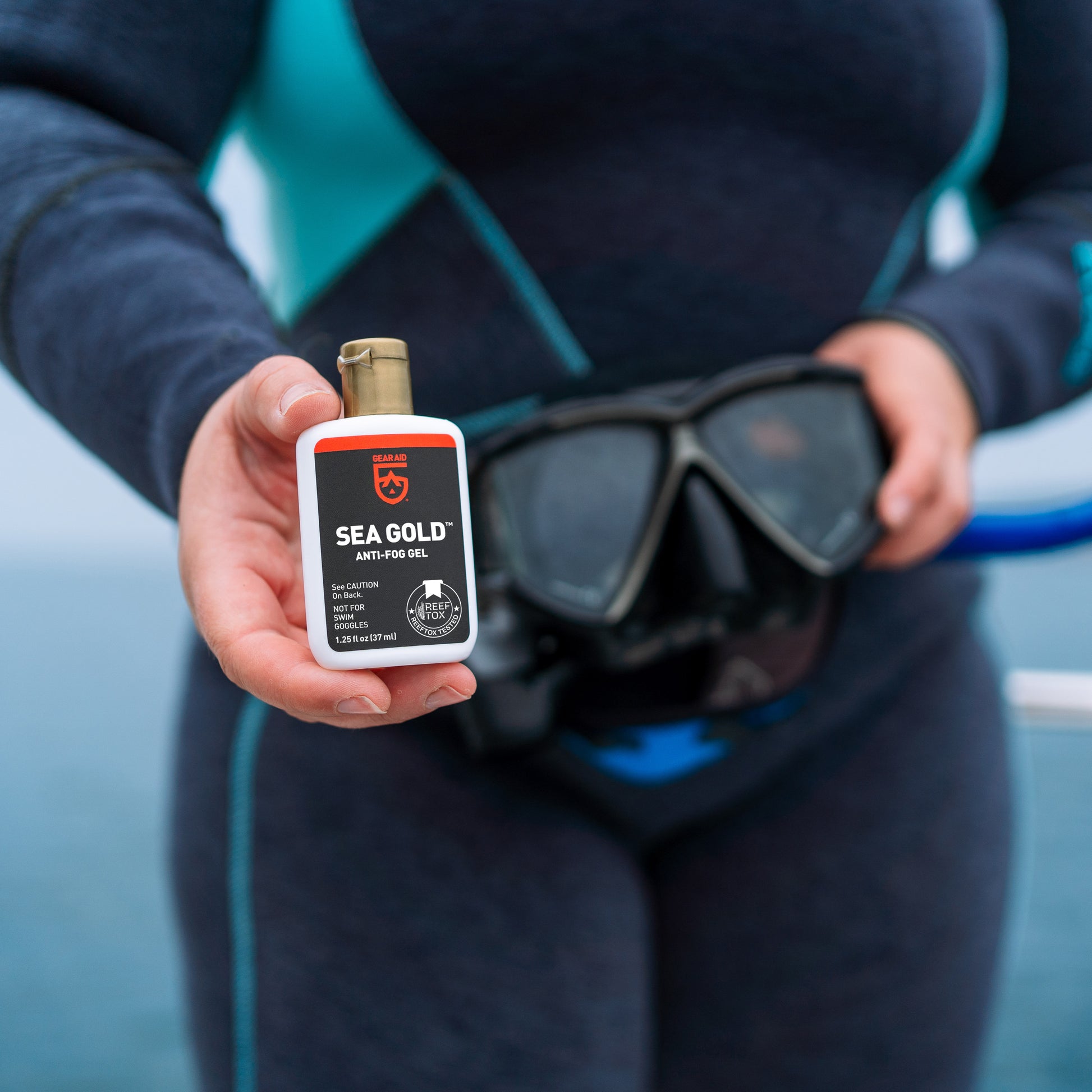 Woman wearing a wetsuit and holding a dive mask in one hand a bottle of Sea Gold Anti-Fog Gel in the other hand